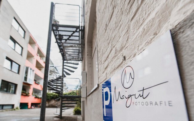 Detailaufnahme eines Maigut Fotografie-Schilds mit Blick auf eine Wendeltreppe in einem Hinterhof in Köln-Ehrenfeld als Teil des visuellen Markenauftritts.