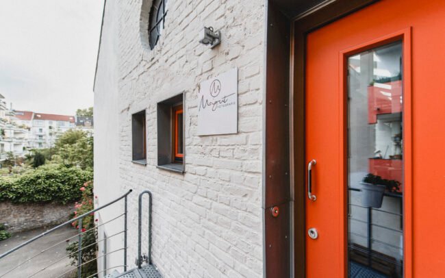 Außenansicht des Agentureingangs von Maigut Media mit roter Tür und Firmenlogo in urbaner Ehrenfelder Umgebung.